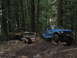 1990 Ford Bronco - Two modified off-road vehicles sit in a wooded area, one appearing burnt and the other a blue buggy.
