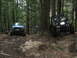 1990 Ford Bronco - Two off-road vehicles navigate a muddy, wooded trail with one vehicle appearing to be winched.