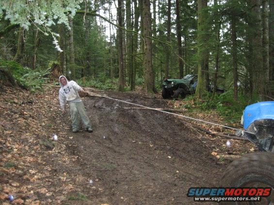 Person pulling a tow rope in a muddy forest with two off-road vehicles visible