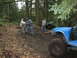1990 Ford Bronco - Two men pull a rope to help a blue off-road vehicle navigate a muddy trail in a wooded area.