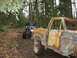 1990 Ford Bronco - Two vehicles navigate a muddy path through a dense forest, with one pickup truck appearing heavily damaged by fire.