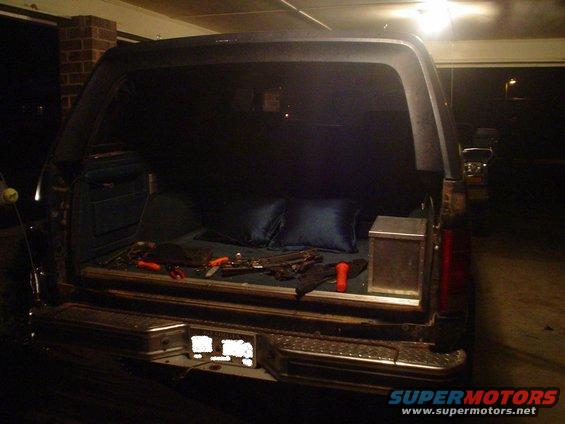 Rear view of an SUV, possibly a Ford Bronco, with the tailgate open. Tools and two blue pillows are visible in the cargo area