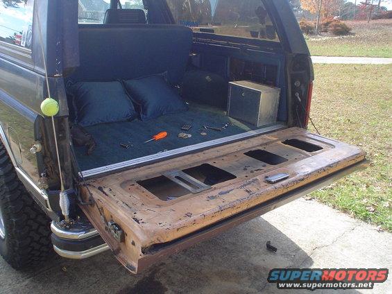Rusty tailgate of a dark blue 1980s Ford Bronco, open, showing interior cargo area with two pillows and tools