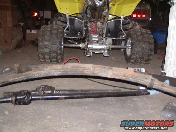 Rear of yellow ATV with driveshaft and leaf spring in foreground. Car taillights visible in background