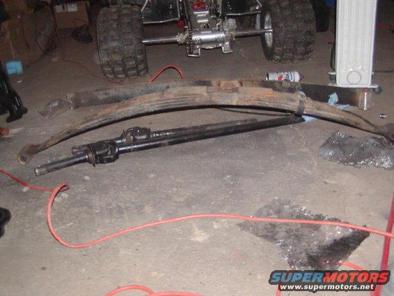 Rusty leaf springs and a driveshaft lie on a concrete floor next to a four-wheeler