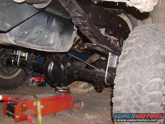 underneath vehicle, rear axle, leaf springs, fuel tank, and tire are visible. A red jack supports the axle