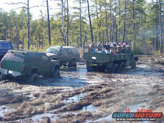 Green SUV being towed by a military truck in a muddy forest clearing