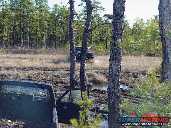 Two trucks stuck in mud, one with door open. Trees and dry grass in background