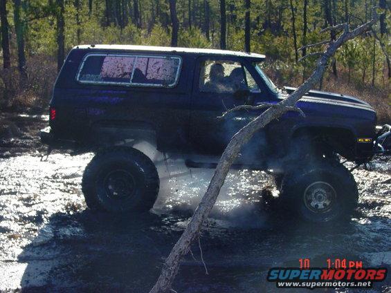 Off-road SUV with large tires stuck in mud, trees in background. Watermark reads SUPERMOTORS