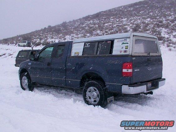Blue Ford F-150 truck with a white camper shell driving in snow, another truck behind it