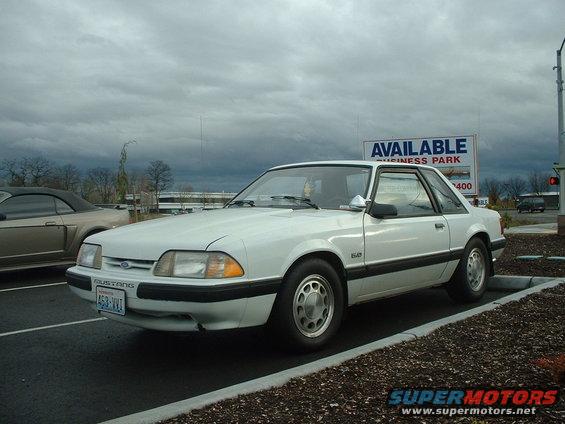 White 1987 Ford Mustang coupe parked next to a tan convertible. Sign says 'Available Business Park'