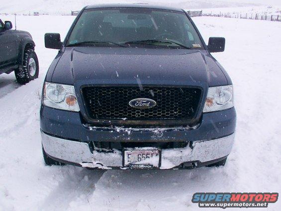Dark blue Ford F-150 truck front grille and bumper covered in snow, license plate E 6557