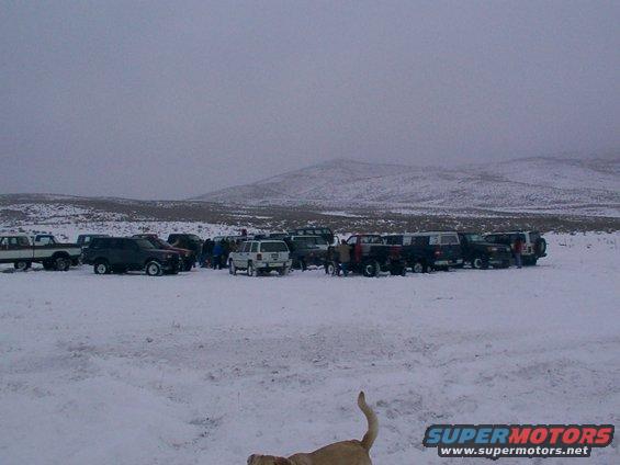 Several SUVs and pickup trucks parked in a snow-covered field with a group of people standing around