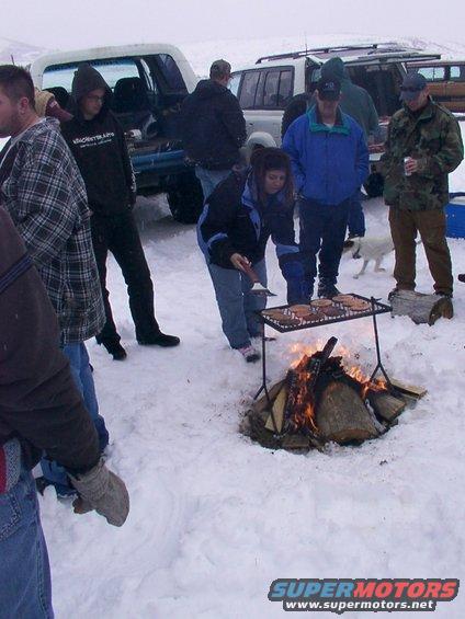 People in snow gathered around a campfire with a grill cooking food. Several SUVs and trucks are parked in the background