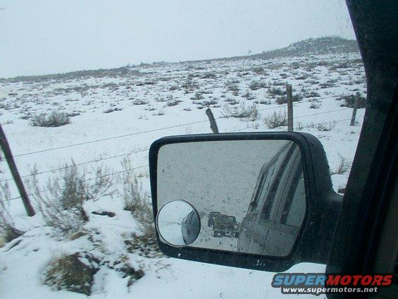 Two trucks drive on a snow-covered road, seen in a side mirror