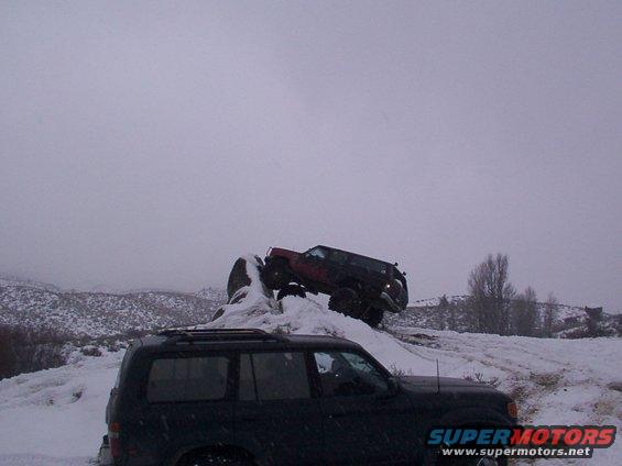 Two SUVs off-roading on a snowy hill; one is dark colored in the foreground, the other is red and climbing a rock