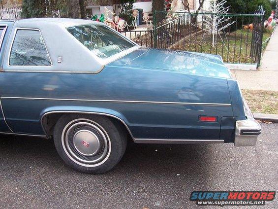 Rear quarter view of a blue sedan with a light-colored vinyl roof and chrome trim. It has a red reflector on the rear fender