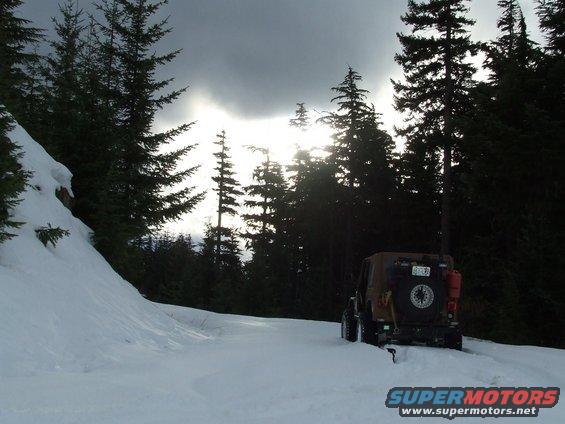 Jeep Wrangler on snowy road in forest, sun breaking through clouds