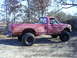 1994 Ford F-250 - A red pickup truck with off-road tires sits on a dirt path surrounded by trees.