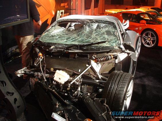 Silver sports car with a cracked windshield, front-end damage, and exposed engine. Two orange sports cars are visible in the background