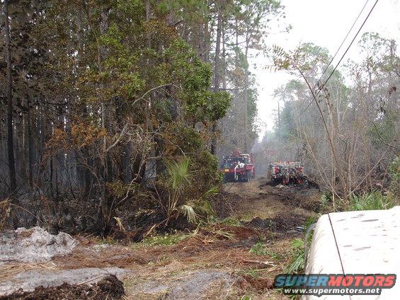 Two red fire trucks on a muddy dirt road through a wooded area with some smoke visible