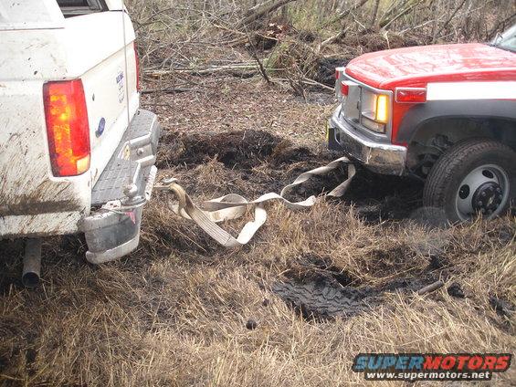 Rear view of a white 1989 Ford Bronco being towed by a red truck stuck in mud