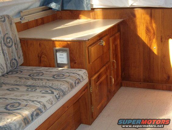 Interior of a pop-up camper showing wood paneling, a patterned seat cushion, and cabinets