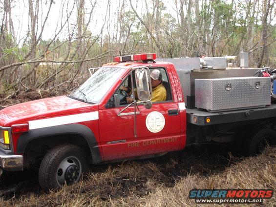 Red fire department truck with a white stripe on the side, driving through muddy terrain with trees in the background