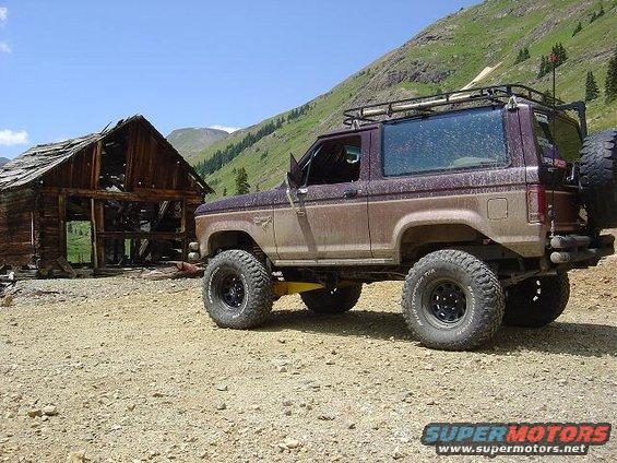 Modified 1986 Ford Bronco II parked next to an old wooden structure in a mountainous area