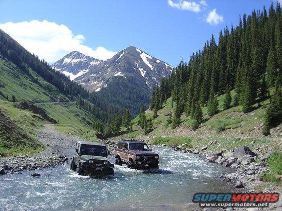 Brown 1986 Ford Bronco II driving through river, mountains and trees in background