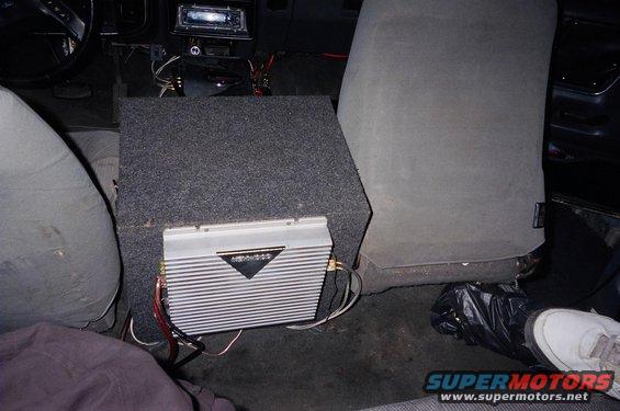 Car interior with a large subwoofer box and amplifier installed behind the passenger seat. Gray fabric seats and dark carpeting are visible