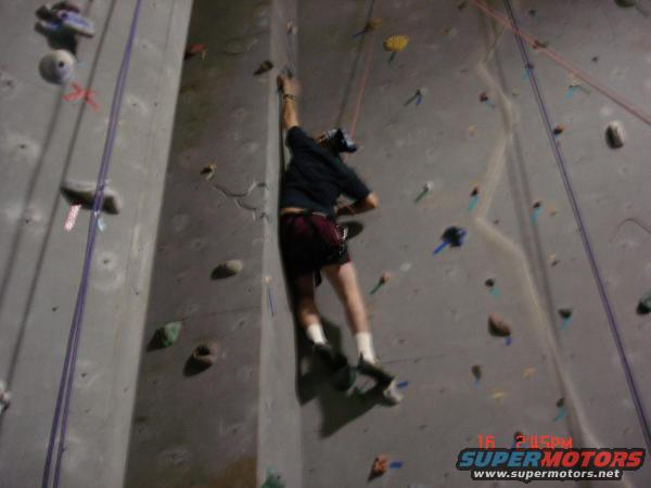 Person in dark shirt and red shorts climbing an indoor rock wall with ropes and colorful holds