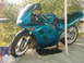 2001 BMW 3 Series - A teal Suzuki motorcycle is parked on a concrete surface next to a white pole and some bushes.