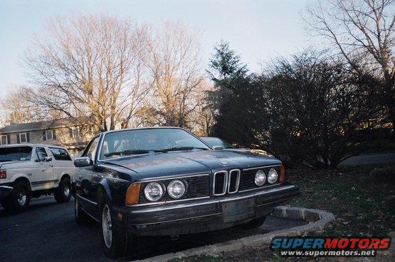 Dark blue BMW coupe parked next to a white SUV. Trees and houses in background