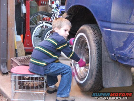 Boy in blue jeans and striped sweater uses a wrench on a car's wheel. Garage background with bikes