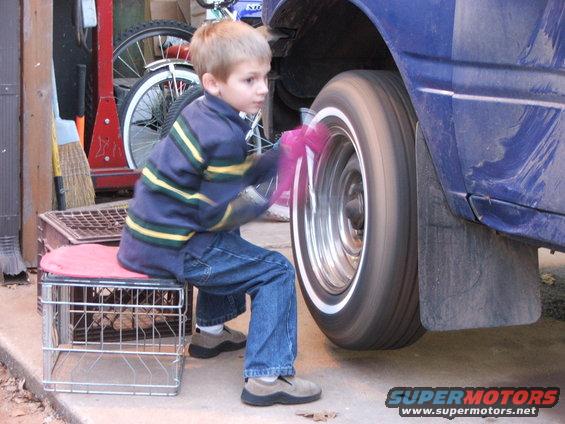 Boy sits on a stool cleaning the spinning wheel of a blue car