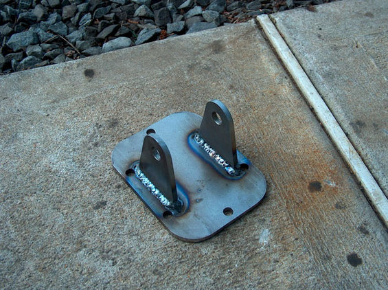 metal bracket with two upright tabs, welded onto a flat base with four bolt holes