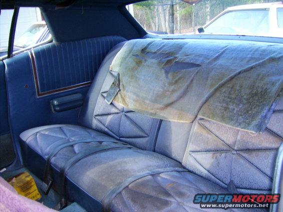 Rear seat of a car with blue upholstery, torn cover on the backrest, and seatbelts