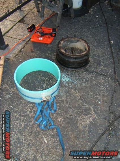 Two automotive parts, a blue pipe section with blue straps and a metal rim with studs, sit on asphalt next to an orange power tool