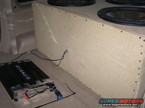 Car interior with a large subwoofer box and an amplifier. Wires connect the components