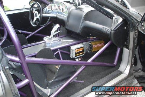 Interior of a race car with purple roll cage, racing seats, and aftermarket shifter. "Supermotors" logo visible