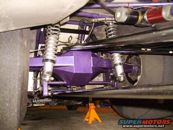 Underside view of a car's rear axle, differential, and suspension system, painted purple. Large tires are visible