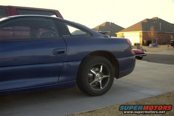 Side view of a blue sports coupe parked on a driveway. Another vehicle is visible in the background