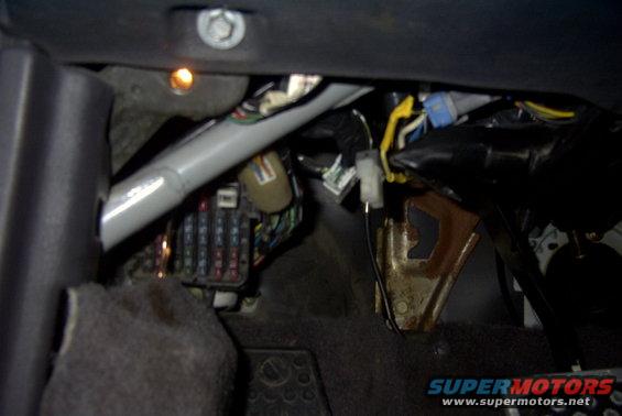 Vehicle interior showing a fuse box, wires, and metal brackets. A small orange light is visible