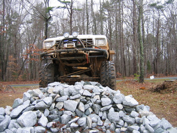 Modified off-road truck with large tires and winch, parked in front of a pile of rocks and trees