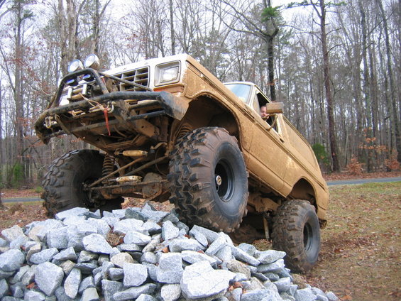Brown off-road SUV with large tires climbing over rocks, front grille and headlights visible