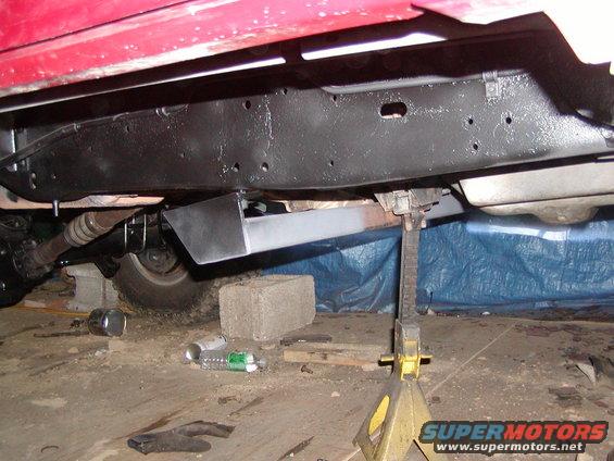 Underside of a red vehicle on jack stands, showing the frame, drive shaft, and a metal plate