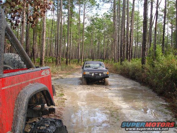 Dark SUV driving through mud, red Jeep in foreground, pine forest
