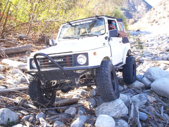 White 1987 Suzuki Samurai with aftermarket bumper and large tires navigating a rocky stream bed