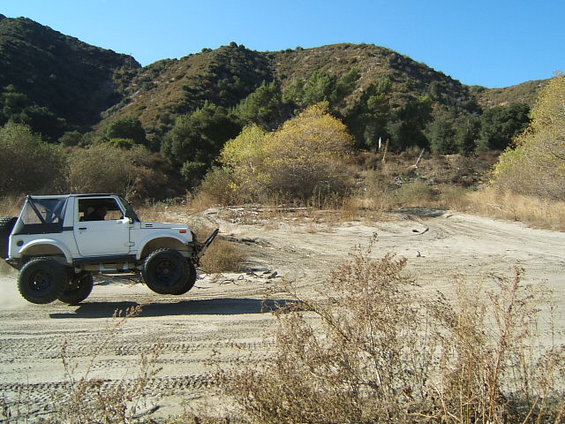 White Suzuki Samurai airborne on a dirt track with hills in the background